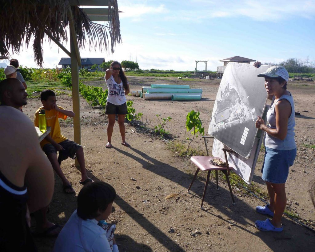 People gathered around a board of an arieal view of the Mokauea Island.